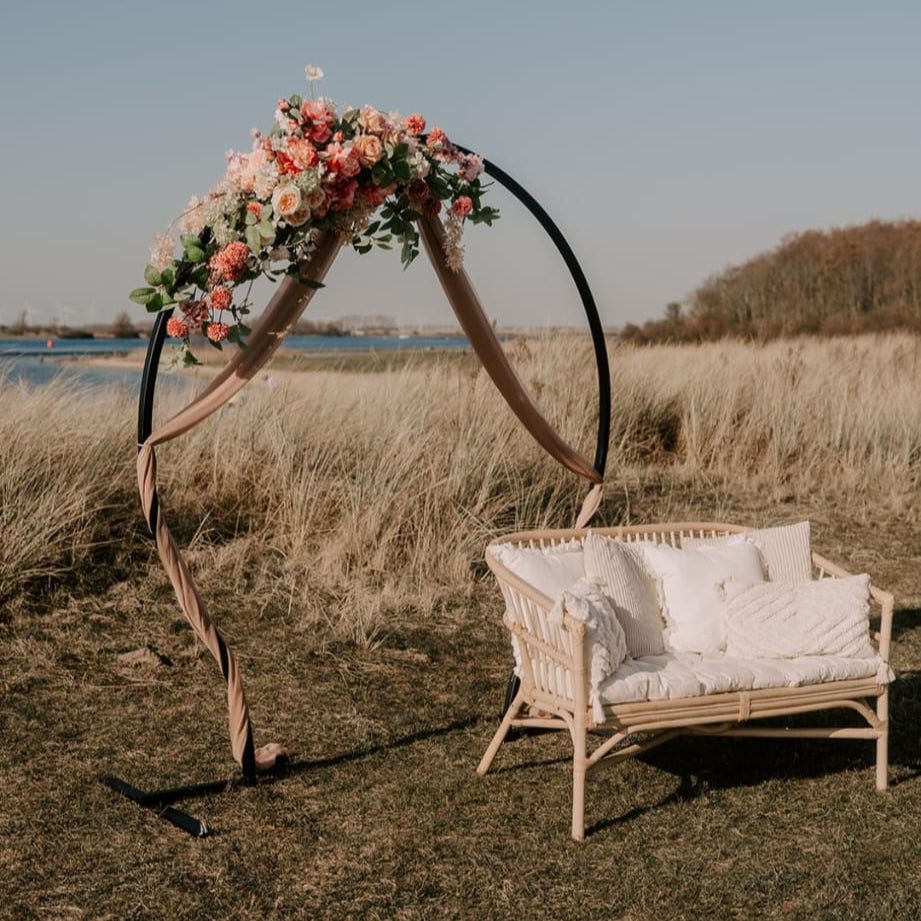 A floral arch with pink and white flowers stands next to a white wicker bench on a grassy field by a body of water.