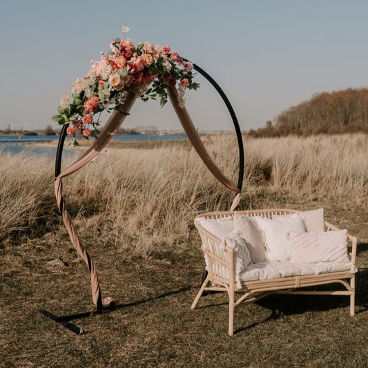 A floral arch with pink and white flowers stands next to a white wicker bench on a grassy field by a body of water.