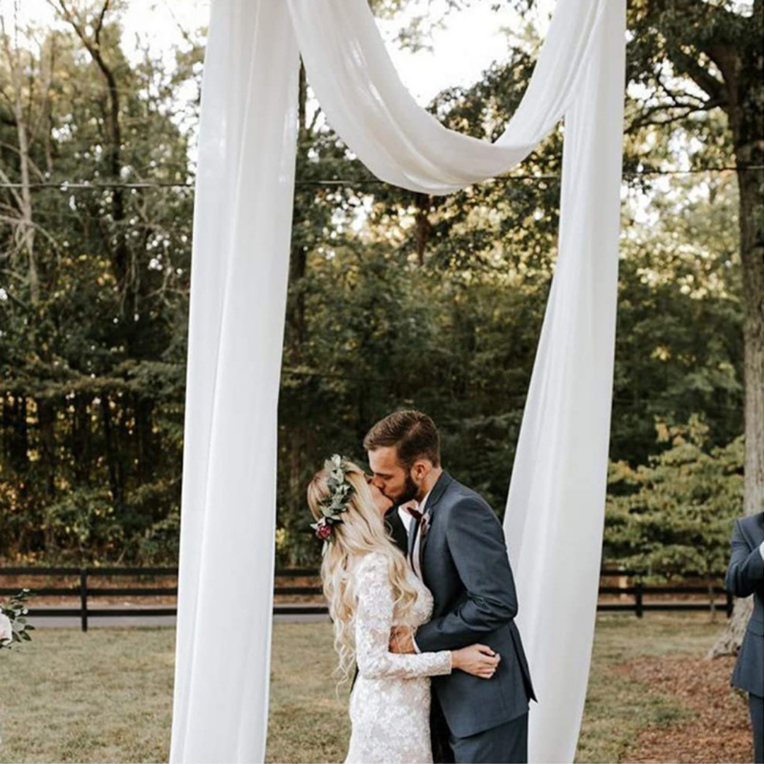 in het bos op een weiland met een hek eromheen staat een boom met 1 grote tak waar wit organza doek over drappeerd, daar onder/tussen staat een zoenend bruidspaar. zij staat links en hij rechts